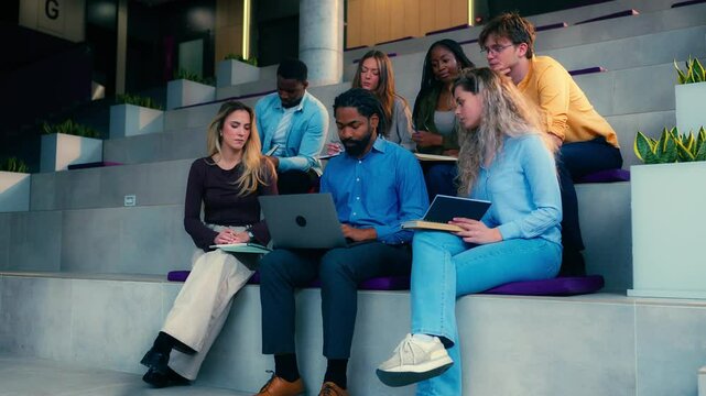 University students are engaging in a collaborative project, utilizing a laptop and notebooks while seated on steps in a modern academic setting, captured in slow motion