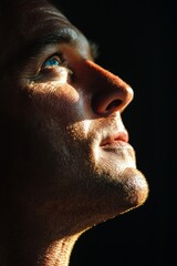 Obraz premium A handsome man, a close-up of his face in the dark, illuminated by sunlight from above. The light shines on his eyes and nose, creating a soft glow effect. He is looking up at something