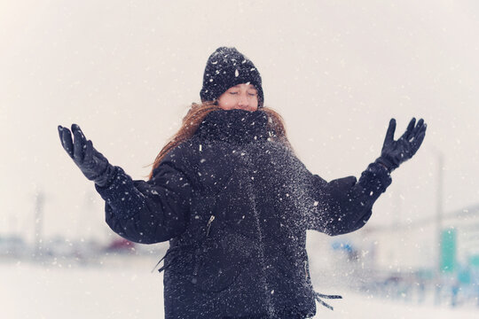 A cute, happy teenage girl in a knitted hat, scarf, and winter clothes joyfully plays under the falling snow. Festive winter mood