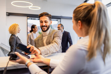 Businessman arriving in a hotel with his colleagues for a business conference, paying with smart phone for room at the reception.