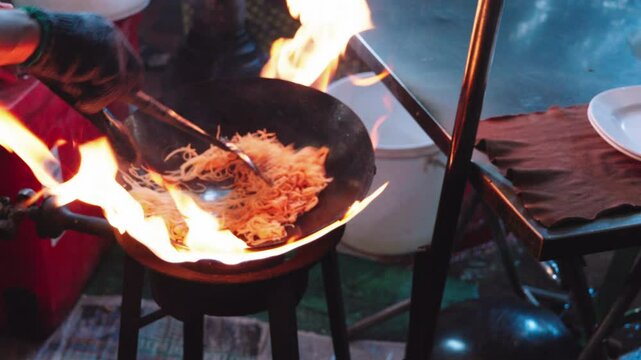 Street vendor chef cooking and throwing stir-fried Pad Thai noodles in a sizzling wok over a roaring open flame at a bustling street food stall in Bangkok's vibrant Chinatown, Thailand