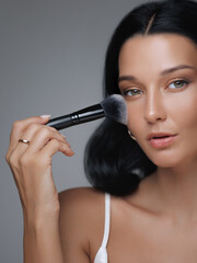 Close-up portrait of a beautiful woman applying makeup with a brush, highlighting skincare and...