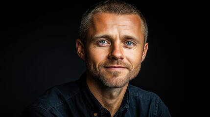 Obraz premium A man is posed against a dark backdrop, showcasing his blue eyes and confident smile. His casual attire highlights a relaxed yet professional demeanor