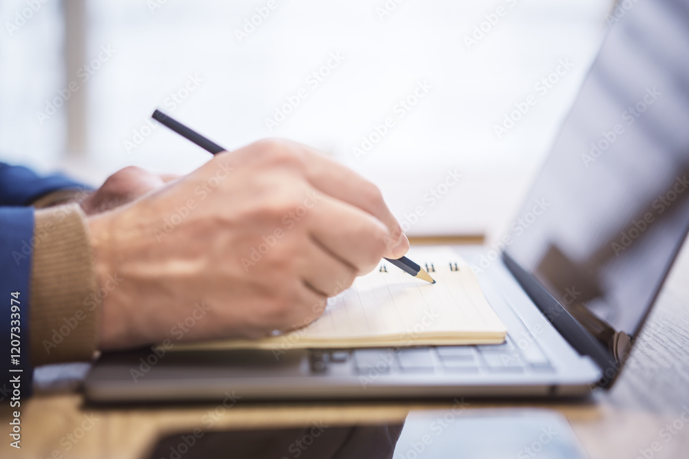 Poster in close view, a man's hand writing down notes in a notepad on a laptop, against a hazy backdrop