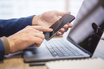 Detailed view of a man utilizing his modern smartphone at his workspace, laptop in the vicinity against the fuzzy backdrop of his busy office environment
