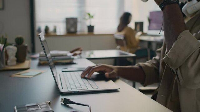 Midsection shot of unrecognisable Black male developer with beard drinking coffee from reusable cup while coding on laptop at work desk