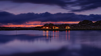 Calm winter morning with the Sun below horizon behind a small island with some houses on, and their reflection on icy lake in foreground. Lofoten Islands, Northern Norway