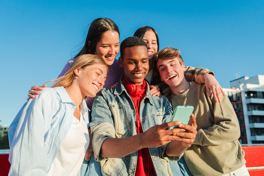 A joyful group of diverse friends enjoying a moment together outdoors, smiling and using a cellphone to capture memories, creating bonds and sharing happiness. Teenage students watching a smart phone