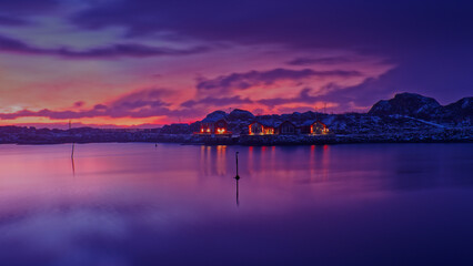 Cloudy reddish pink sky with sunrise behind a small island with some houses on and their reflection on calm lake in foreground. Lofoten Islands, Northern Norway.