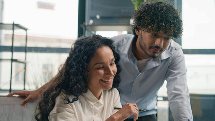 Indian businessman mentor coach help Arabian businesswoman with laptop teaching help explain online project paperwork two smiling multiracial woman and man business colleagues cooperate work in office