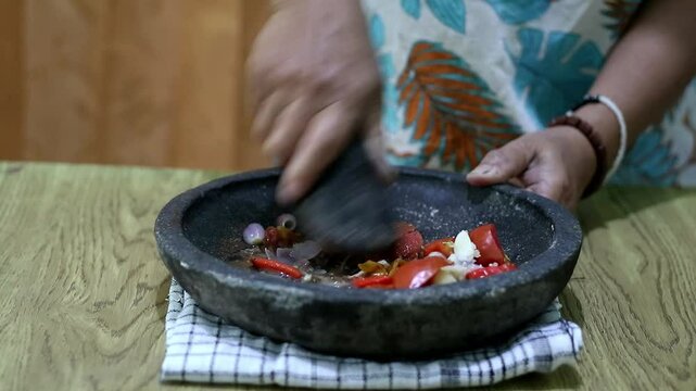 Indonesian woman's hands grinding spices on a stone mortar for sambal. Traditional Indonesian sambal being prepared with mortar and pestle. Asian womans hands crushing chili peppers and spices 