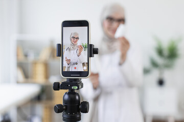 Muslim woman in hijab demonstrating medicine bottles while recording video. Young adult female creating educational content on healthcare. Professional setting suggests engaging online audience.
