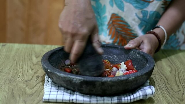 Indonesian woman's hands grinding spices on a stone mortar for sambal. Traditional Indonesian sambal being prepared with mortar and pestle. Asian womans hands crushing chili peppers and spices 