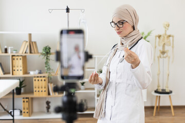 Muslim woman doctor demonstrates medical procedure in online video. Focus on education and healthcare. She is wearing glasses and hijab while explaining methods to her audience.