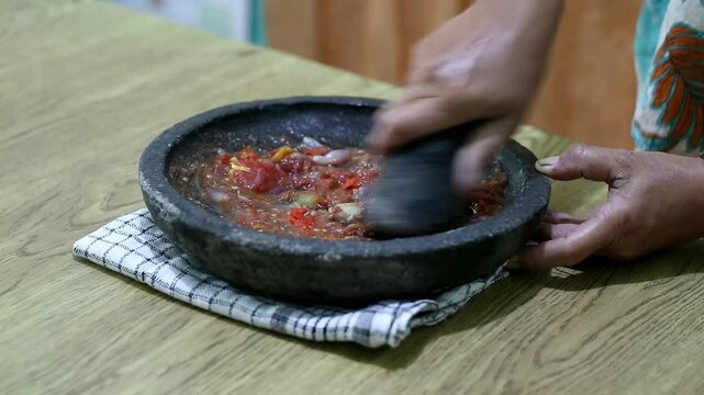 Indonesian woman's hands grinding spices on a stone mortar for sambal. Traditional Indonesian sambal being prepared with mortar and pestle. Asian womans hands crushing chili peppers and spices 