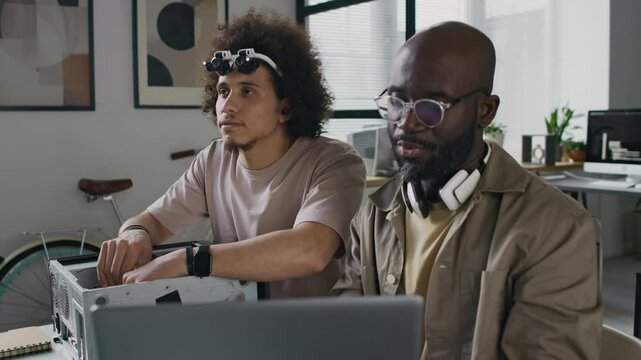 Millennial male web engineer pointing at laptop screen while gen Z repairman with magnifying goggles putting hard disk drive in computer case at workplace