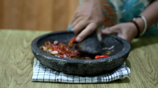 Indonesian woman's hands grinding spices on a stone mortar for sambal. Traditional Indonesian sambal being prepared with mortar and pestle. Asian womans hands crushing chili peppers and spices 