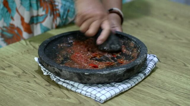 Indonesian woman's hands grinding spices on a stone mortar for sambal. Traditional Indonesian sambal being prepared with mortar and pestle. Asian womans hands crushing chili peppers and spices 