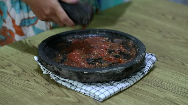 Indonesian woman's hands grinding spices on a stone mortar for sambal. Traditional Indonesian sambal being prepared with mortar and pestle. Asian womans hands crushing chili peppers and spices 