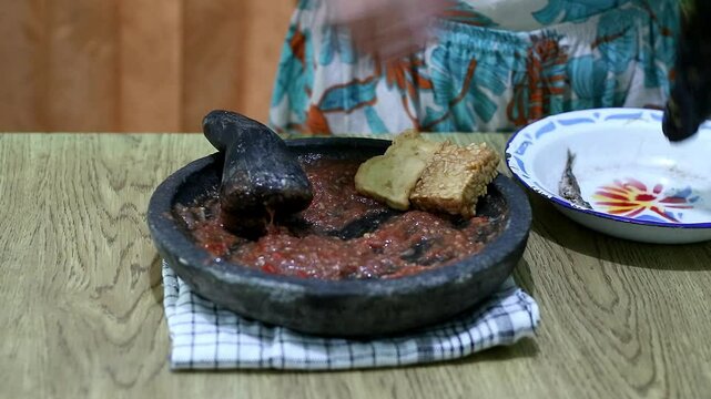 Indonesian woman's hands grinding spices on a stone mortar for sambal. Traditional Indonesian sambal being prepared with mortar and pestle. Asian womans hands crushing chili peppers and spices 