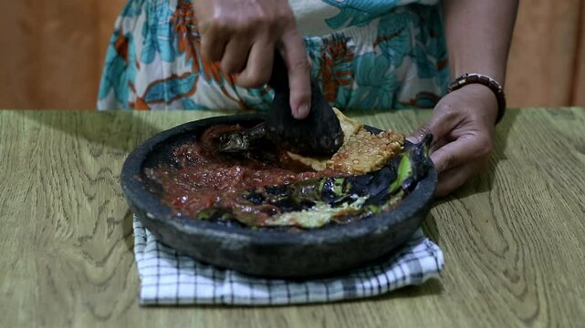 Indonesian woman's hands grinding spices on a stone mortar for sambal. Traditional Indonesian sambal being prepared with mortar and pestle. Asian womans hands crushing chili peppers and spices 