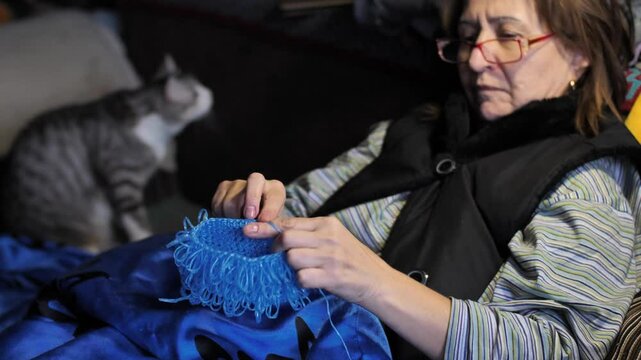 Woman crocheting a product made of blue polypropylene nylon plastic twine, sitting on the couch. A cat is playing with a phone nearby