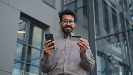 Happy Indian Arabian businessman use credit card online bank payment in city near office business customer entrepreneur man with mobile phone internet shopping buying easy pay money banking service