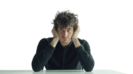 A frustrated young man sitting at a table, holding his head in his hands, expressing feelings of stress and overwhelm against a white isolated background.