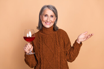Charming elderly woman holding a wine glass with a friendly smile against a beige backdrop