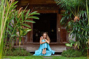 Young woman sit on open-air veranda of wooden bungalow with tropical garden view. Girl relaxing in...