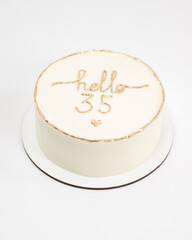 A beautifully decorated small cake, off-white frosting with gold lettering that says 'Hello 35' sits on a small white cake board. A simple yet elegant design.