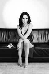 Fototapeta premium A stylish woman in a dark dress sits gracefully on a black leather sofa. A small bouquet rests beside her. The image is in black and white.