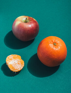 Red apple, orange and half a peeled tangerine on a green background with hard shadow