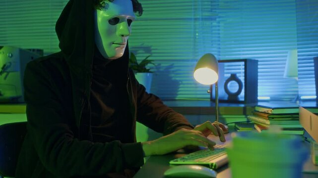 Tilt down shot of male cyber thief with hacker mask maliciously intruding into computer in corporate office in dim light