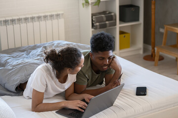 A couple lying on a bed, using a laptop together, engaged in a conversation, with a smartphone placed nearby, in a cozy and relaxed bedroom setting