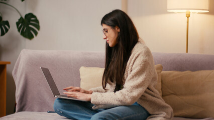Hispanic woman types on laptop sitting on couch at home