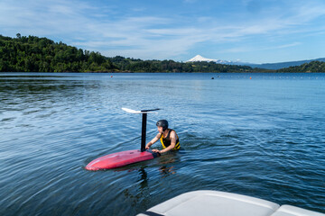 A teenager wearing a life jacket and helmet, floating in the lake alongside a foil board, getting...
