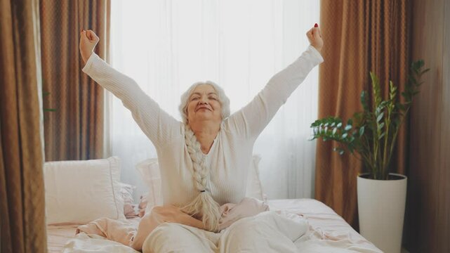 Happy senior elderly woman stretching in bed after waking up, good sleep in cozy room, soft pillow orthopedic mattress cotton fabric bed linen. morning bedroom sun light from window. lady old age 
