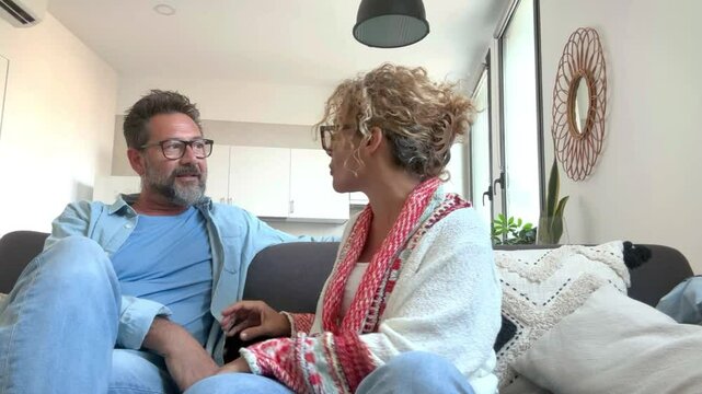 A couple in their 50s, both wearing glasses, sit on a sofa in their living room, enjoying a peaceful Sunday afternoon, chatting calmly and sharing the warmth of their cozy home with white walls.