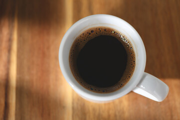 Dark coffee in a white mug resting on a wooden table with soft lighting in the morning