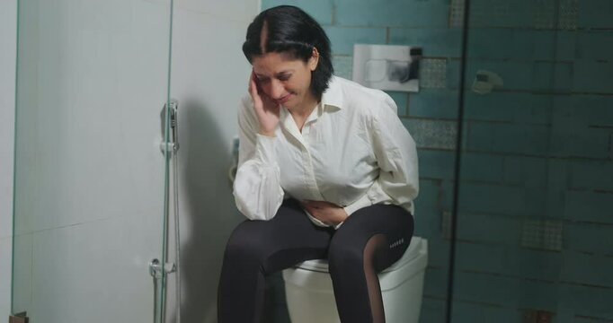 Indian Asian alone older elder mature adult upset sad female woman lady sitting on toilet bowl hold belly waist have health issues feel sick hurt pain ache eat bad food unwell body care indoor home