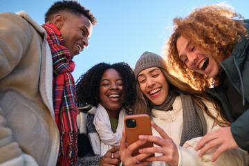 Four cheerful young students are using a smartphone and laughing together during winter holidays