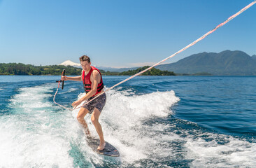 A young man riding a wakesurf board on a bright sunny day in Chile, gripping a tow rope as he glides across the blue lake, with a majestic volcano rising in the background.