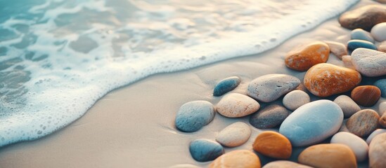 Obraz premium High angle view of colorful smooth stones in shades of blue, orange, and gray on white sandy beach, gentle ocean waves creating tranquil atmosphere.