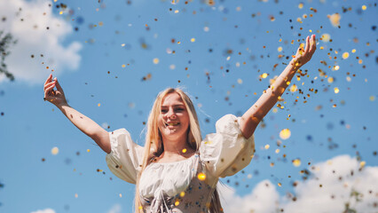 Young blonde woman throwing golden confetti in the air, celebrating success with arms raised and a cheerful smile under a blue sky