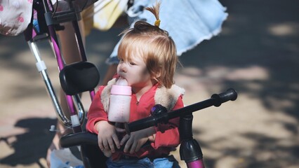 Obraz premium Smiling toddler sipping bottle while riding colorful tricycle on sunny park pathway, childhood joy radiating