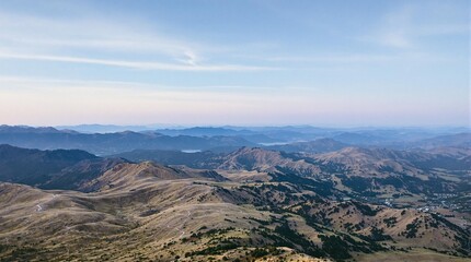 a stunning aerial view of a mountainous landscape with rolling hills and a distant lake under a partly cloudy sky. The land appears rugged and natural, with varying elevations creating a picturesque s