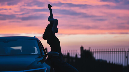 Silhouette of a young woman dancing and enjoying the sunset near her car, expressing freedom and a...
