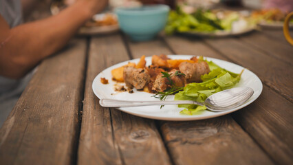 Leftovers of sausages, potatoes and salad on white plate with spoon on wooden table after eating outside