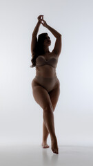 Woman standing in beige lingerie, arms raised gracefully, embodying elegance, natural beauty, and self-love against white background. Silhouette. Concept of natural beauty, self love, confidence.
